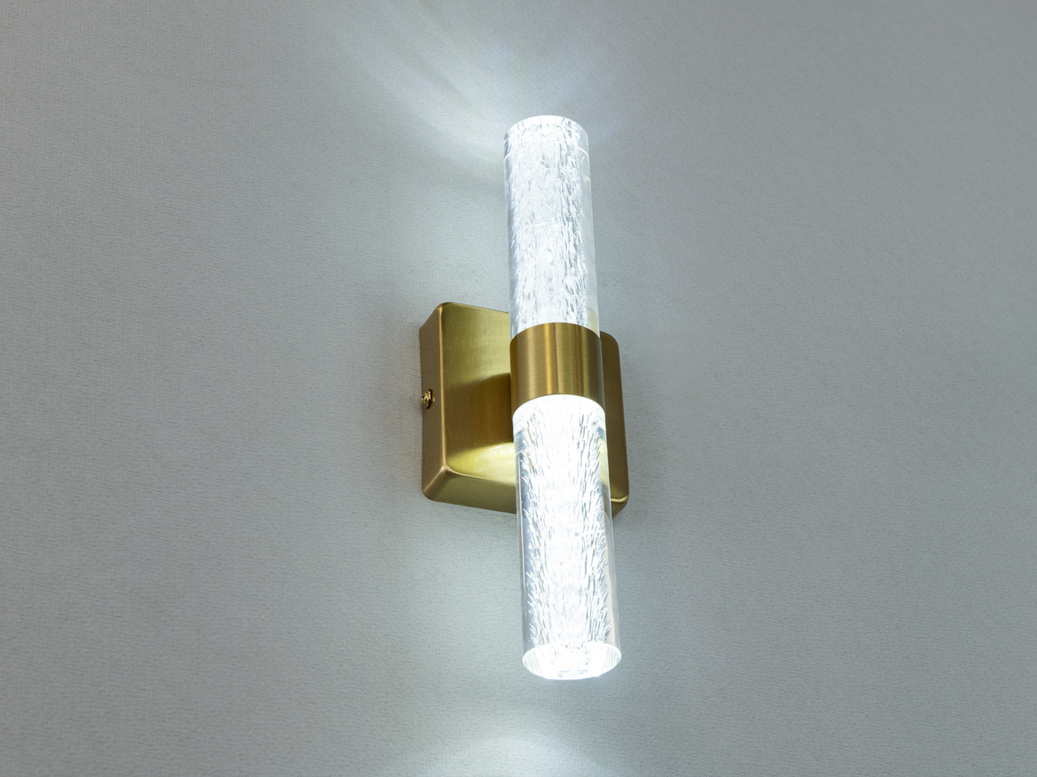 Настінний світильник з латунною основою та текстурованою прозорою трубкою, що забезпечує декоративне освітлення.