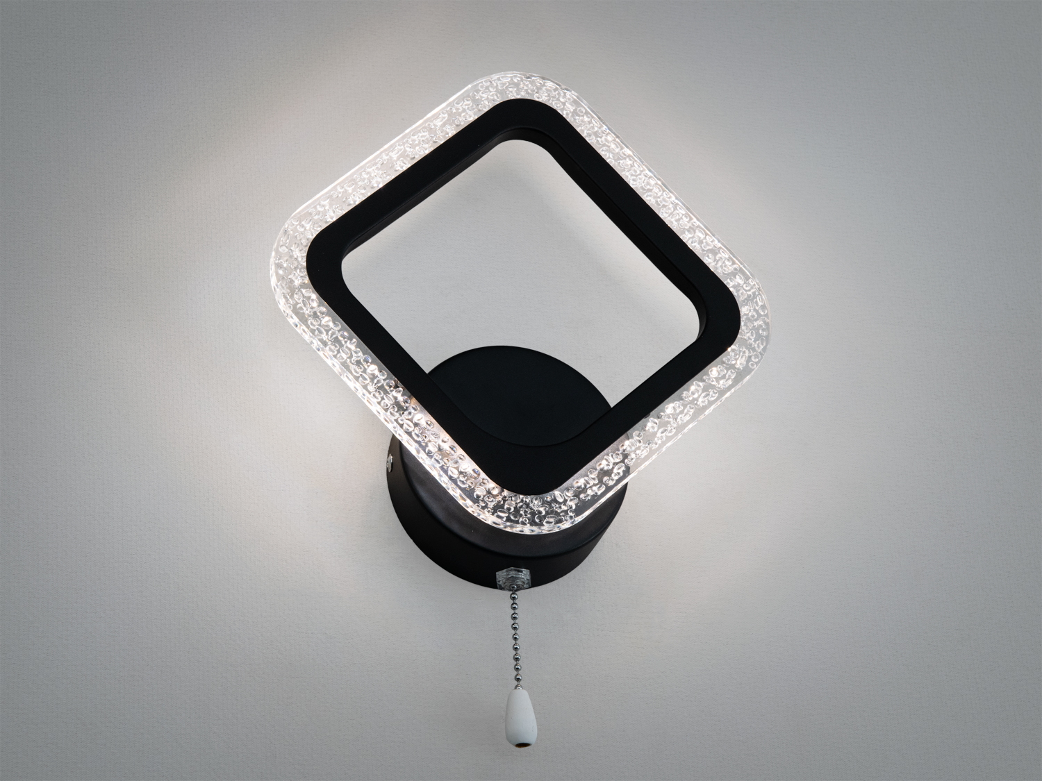 Настінний світильник з квадратною рамкою, що світиться, чорного кольору. Акрилові прозорі вставки створюють м′яке освітлення.