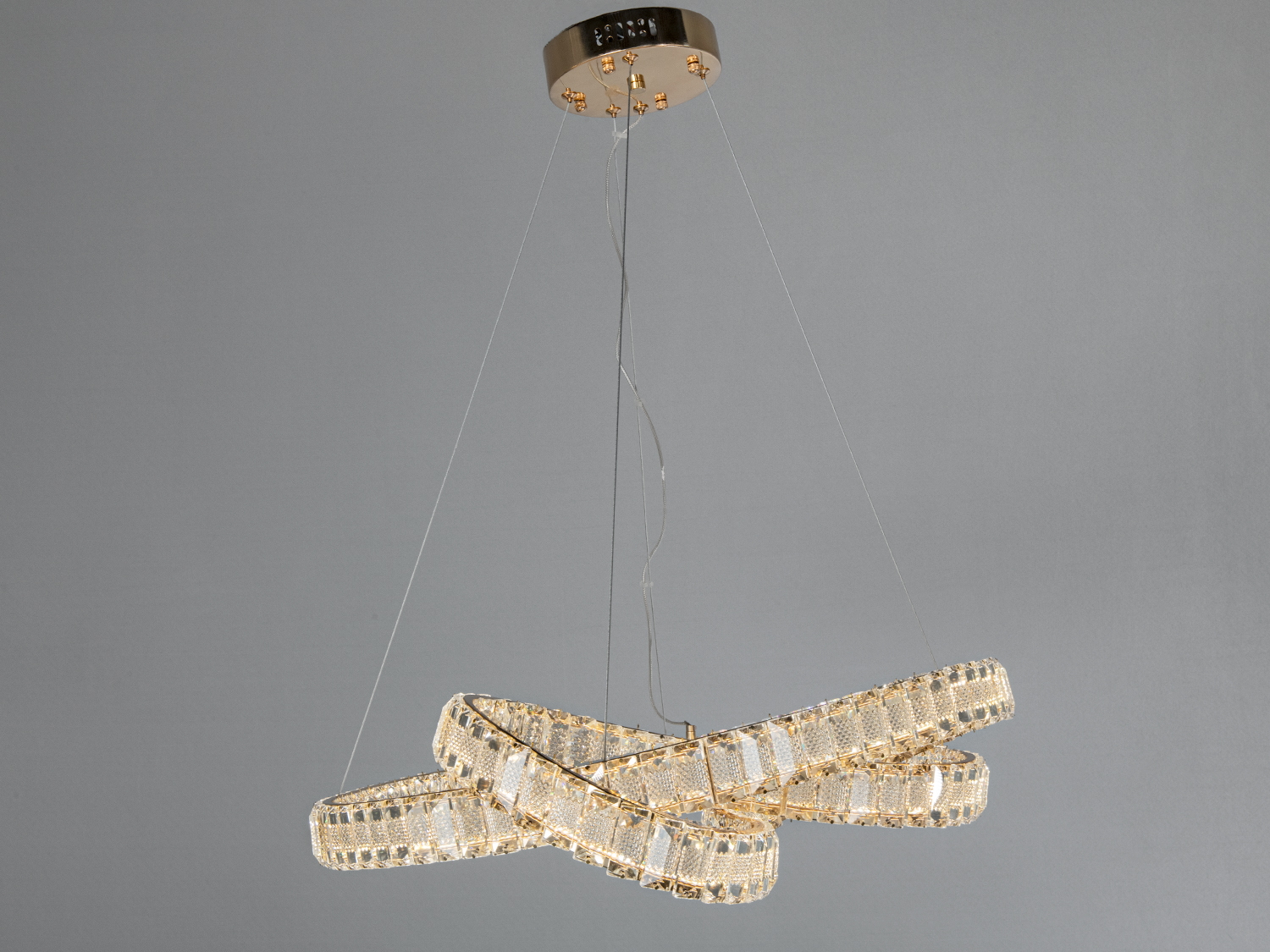 Люстра з кристалами у вигляді переплетених кілець, підвішена на тонких тросах, має металеву основу для монтажу на стелі.